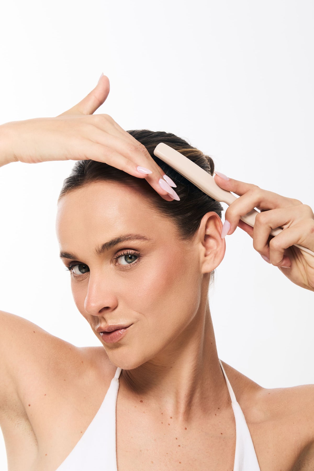 Mulher penteando o cabelo para trás com uma escova, deixando os fios bem alinhados. Ela está usando uma blusa branca de alças finas e aparece em um fundo branco, com o rosto voltado para a câmera.